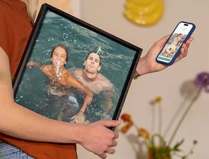 En kvinne holder mobilen sin med fotoknudsen-editoren åpen, mens hun har ferie-veggdekor under armen med to menn som har det gøy i et basseng, som inspirasjon til å gjøre feriebilder om til flott veggkunst.